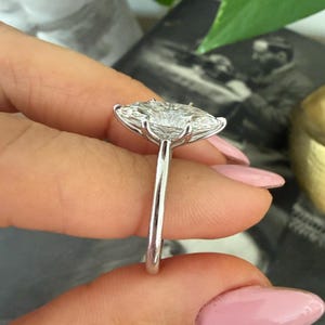 Pu&ograve; includere: Primo piano di un anello d'argento con una grande gemma trasparente taglio a cuscino. La fascia dell'anello &egrave; sottile e lucida, e la gemma &egrave; incastonata con pi&ugrave; griffe. L'anello &egrave; tenuto da dita con smalto rosa.