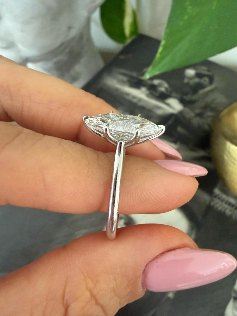 Pu&ograve; includere: Primo piano di un anello d'argento con una grande gemma trasparente taglio a cuscino. La fascia dell'anello &egrave; sottile e lucida, e la gemma &egrave; incastonata con pi&ugrave; griffe. L'anello &egrave; tenuto da dita con smalto rosa.