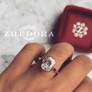 May include: A silver ring with a large, clear, emerald-cut gemstone. The ring is on a person's finger.