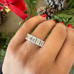 May include: A silver band ring with a row of emerald-cut diamonds. The ring is on a person's finger.
