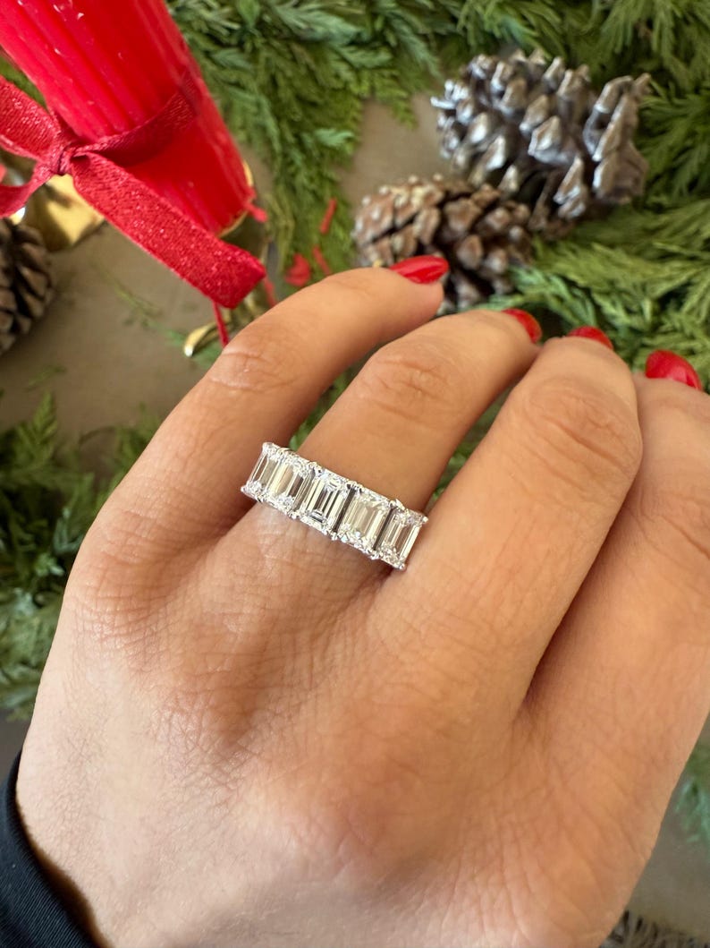 May include: A silver band ring with a row of emerald-cut diamonds. The ring is on a person's finger.