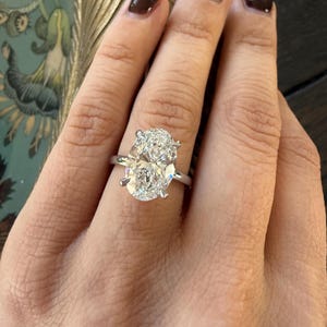 May include: A silver ring featuring a large, oval-cut diamond. The diamond is the focal point, with a brilliant, clear appearance. The ring is set on a slender band, showcasing the diamond's size and sparkle. The ring is a piece of fine jewelry.