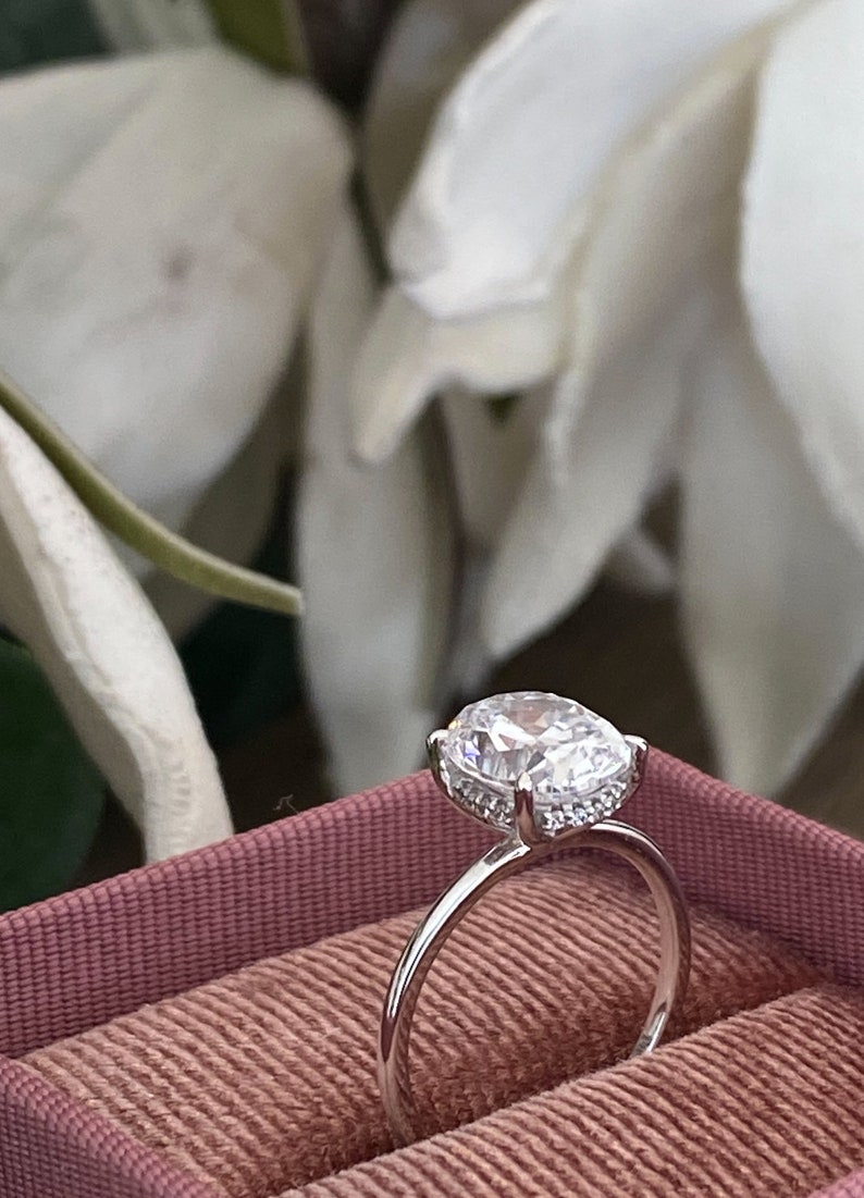 Puede incluir: Un anillo de plata con una gran piedra preciosa redonda y transparente, rodeada por un halo de piedras preciosas m&aacute;s peque&ntilde;as. El anillo est&aacute; en una caja de anillo de terciopelo rosa.