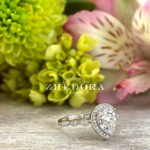 May include: A pear-shaped diamond engagement ring with a halo setting and a pave band. The ring is set in white gold and features a delicate floral design.
