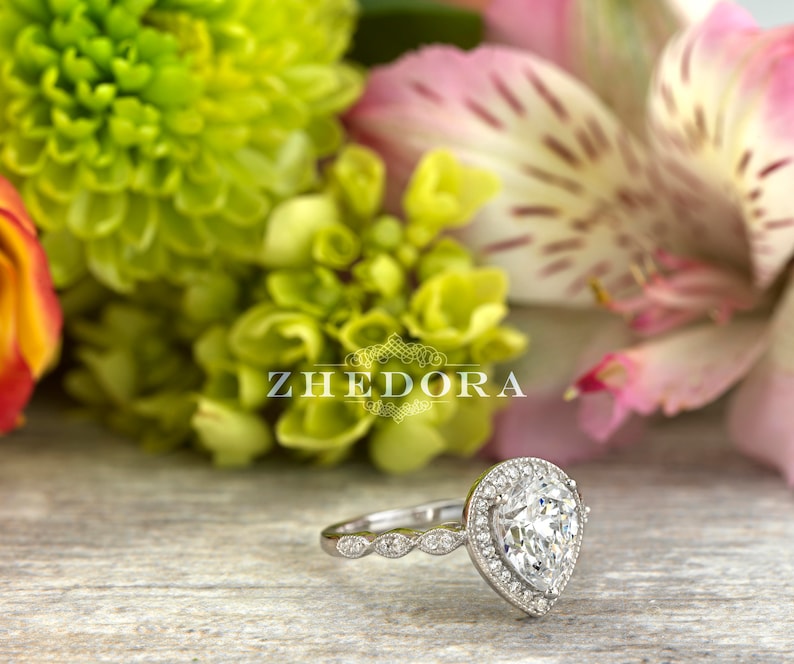May include: A pear-shaped diamond engagement ring with a halo setting and a pave band. The ring is set in white gold and features a delicate floral design.
