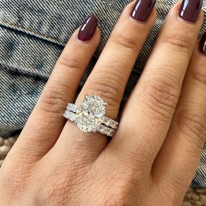 K&ouml;nnte beinhalten: Ein Diamant-Verlobungsring mit einem oval geschliffenen Mittelstein und einem Diamantband. Der Ring befindet sich an einer Hand mit burgunderfarbenem Nagellack, vor einem Jeanshintergrund. Ein Symbol der Liebe.