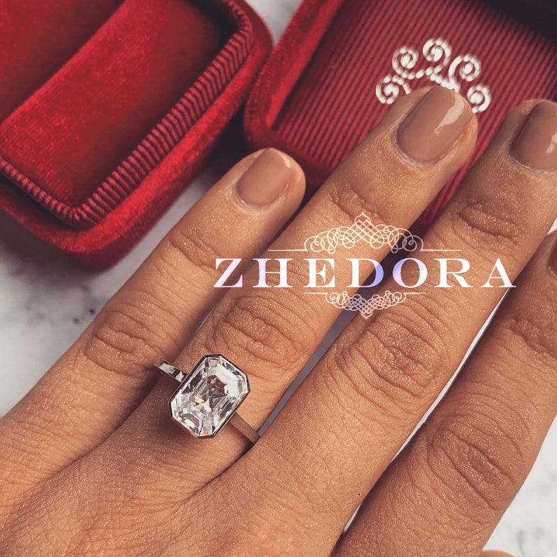 May include: A silver ring with a large, clear, emerald-cut gemstone. The ring is on a person's finger, and the background is a red velvet box.