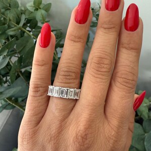 May include: A silver band ring featuring a row of emerald-cut diamonds. The ring is worn on a finger with red nail polish.