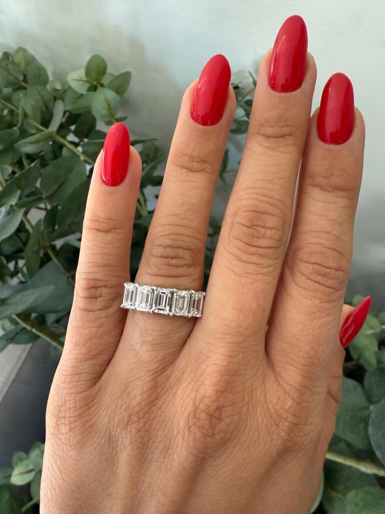 May include: A silver band ring featuring a row of emerald-cut diamonds. The ring is worn on a finger with red nail polish.