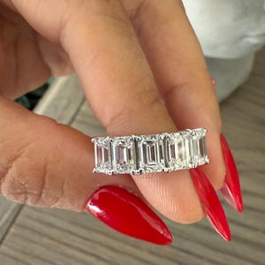May include: A silver ring featuring a row of emerald-cut diamonds. The ring is worn on a finger with red nail polish.