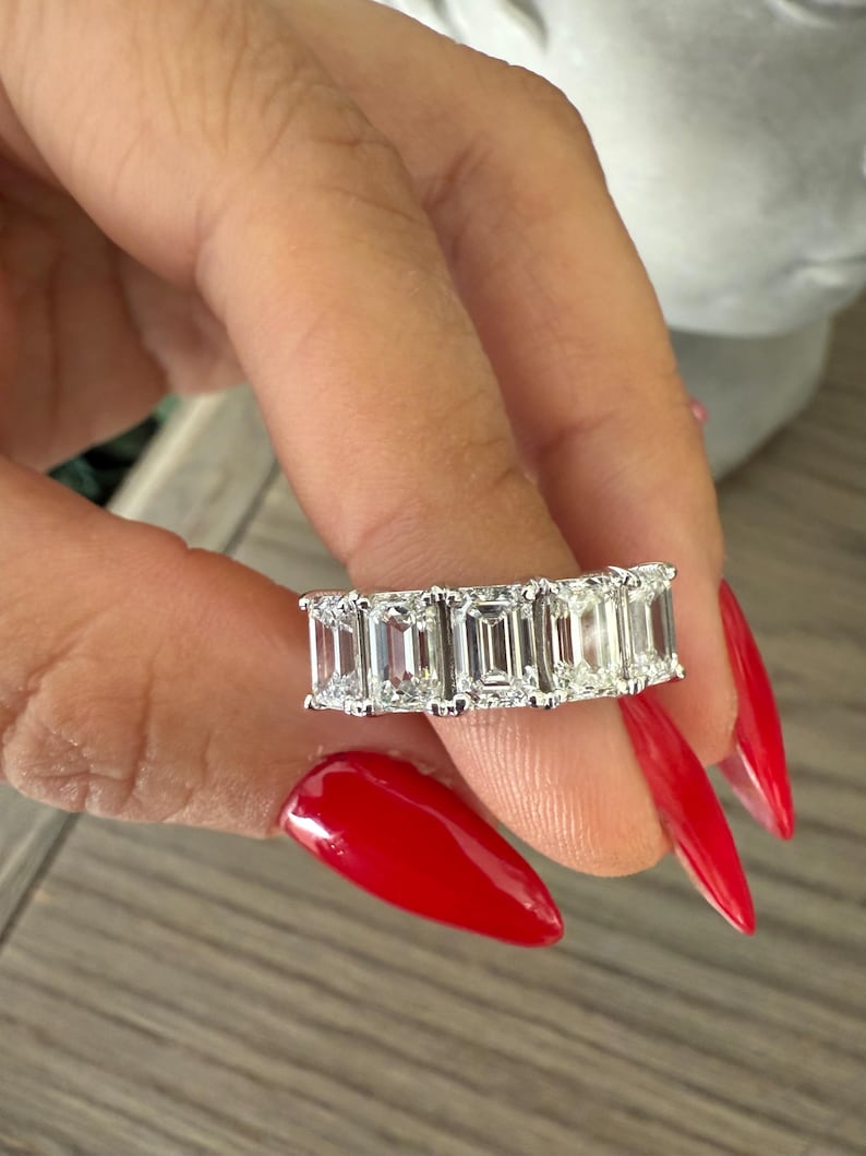 May include: A silver ring featuring a row of emerald-cut diamonds. The ring is worn on a finger with red nail polish.