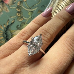 Pu&ograve; includere: Anello di fidanzamento con diamante taglio marquise e fascia argentata. Il diamante &egrave; grande e trasparente, riflettendo la luce. L'anello &egrave; indossato su un dito con smalto rosa chiaro.