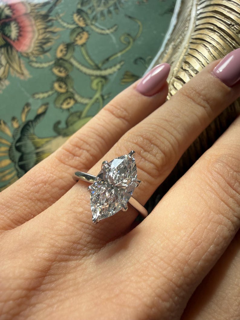 Pu&ograve; includere: Anello di fidanzamento con diamante taglio marquise e fascia argentata. Il diamante &egrave; grande e trasparente, riflettendo la luce. L'anello &egrave; indossato su un dito con smalto rosa chiaro.