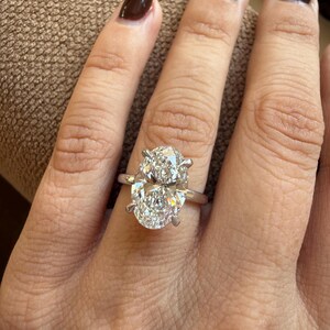May include: A silver ring with a large, oval-cut diamond. The diamond is set with four prongs and sparkles brightly. The ring is on a person's finger, with brown nail polish.