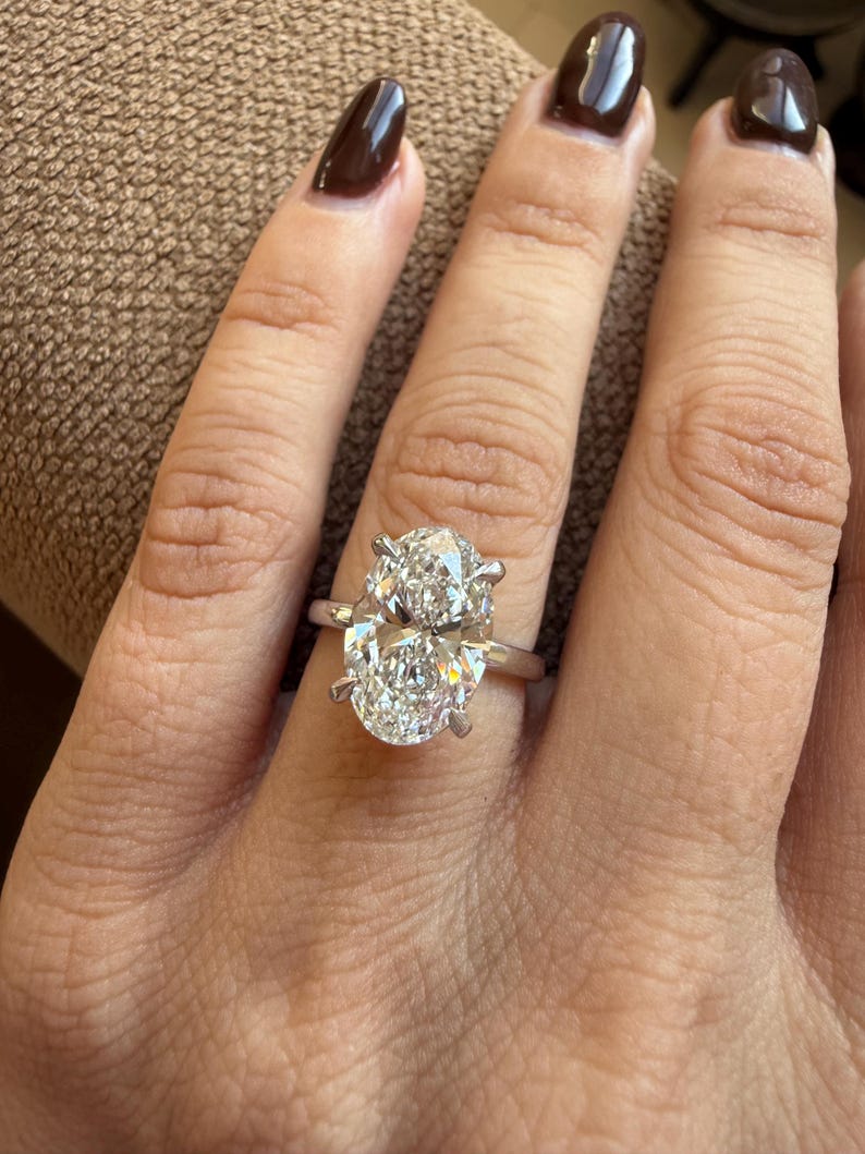 May include: A silver ring with a large, oval-cut diamond. The diamond is set with four prongs and sparkles brightly. The ring is on a person's finger, with brown nail polish.