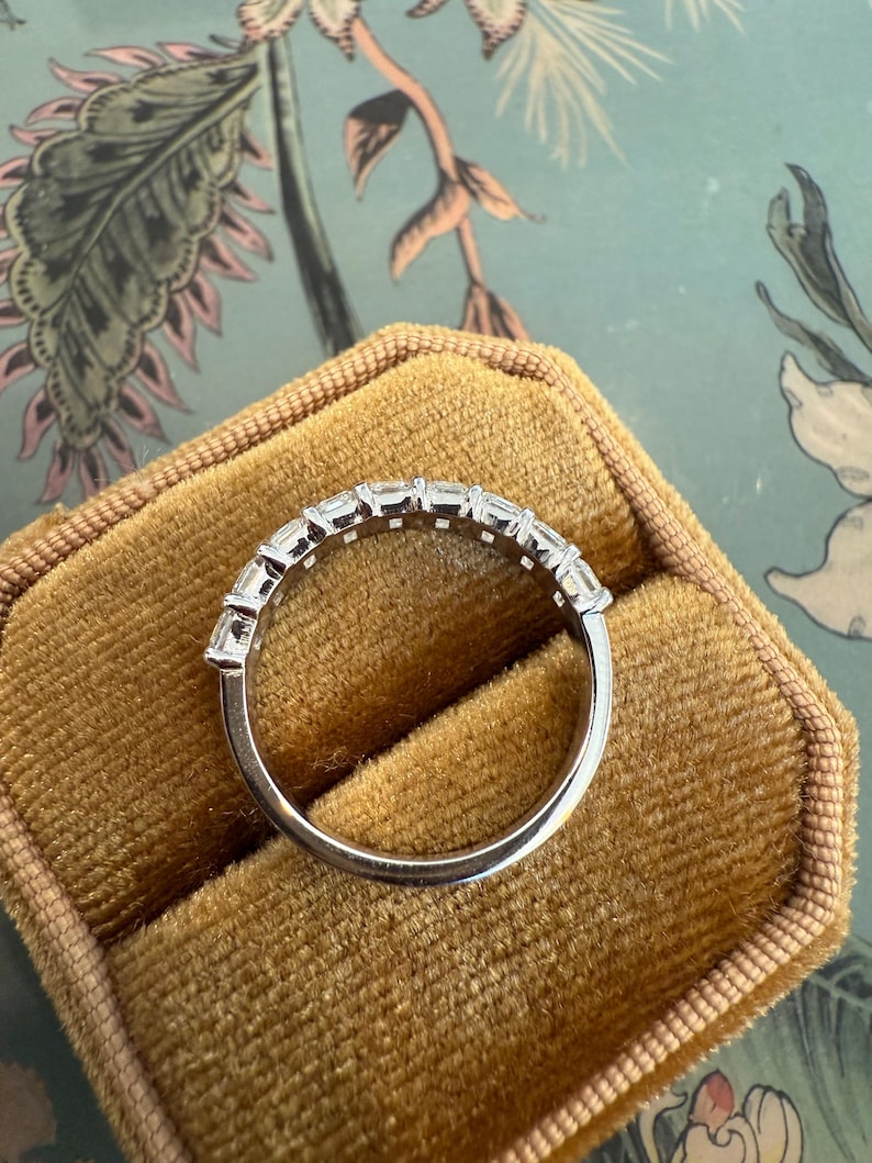 May include: A silver band with a row of small, clear, rectangular-shaped stones set in a continuous line around the band.