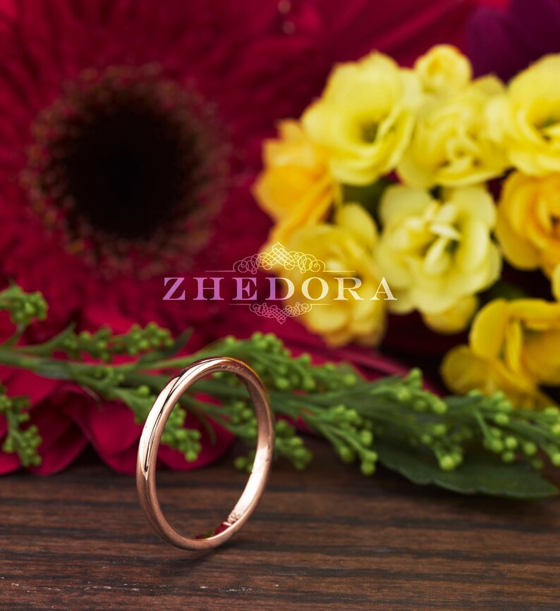 May include: A thin rose gold band ring, lying on a wooden surface with a blurred background of red and yellow flowers.
