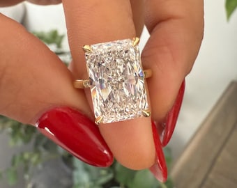 Può includere: Un anello d'oro con un grande diamante taglio radiant rettangolare. Il diamante è tenuto da quattro griffe. L'anello è tenuto da una mano con smalto rosso. Un gioiello.