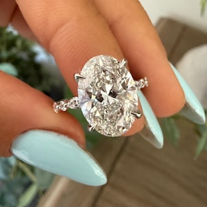 May include: A sparkling engagement ring featuring a large, oval-cut diamond. The diamond is set in a silver band with smaller accent stones. The ring is held up against a light blue background.