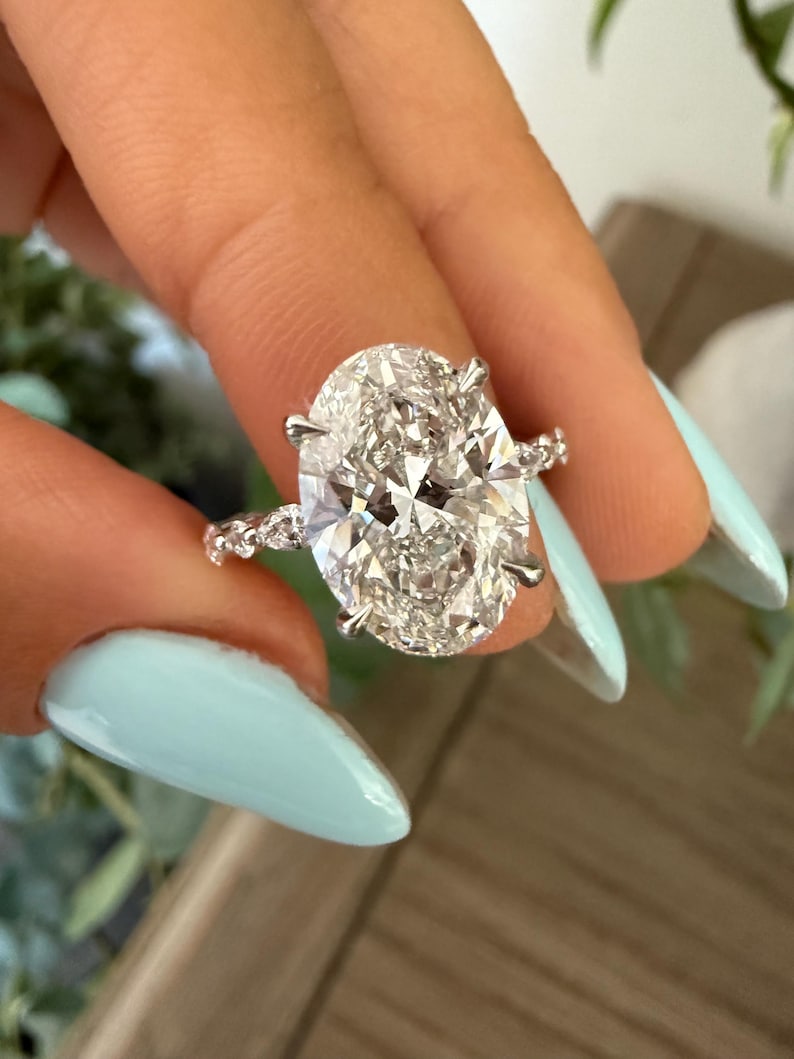 May include: A sparkling engagement ring featuring a large, oval-cut diamond. The diamond is set in a silver band with smaller accent stones. The ring is held up against a light blue background.
