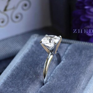 May include: A silver ring with a clear, square-shaped gemstone in a blue velvet ring box. The box has the word "ZHEDORA" printed on it.