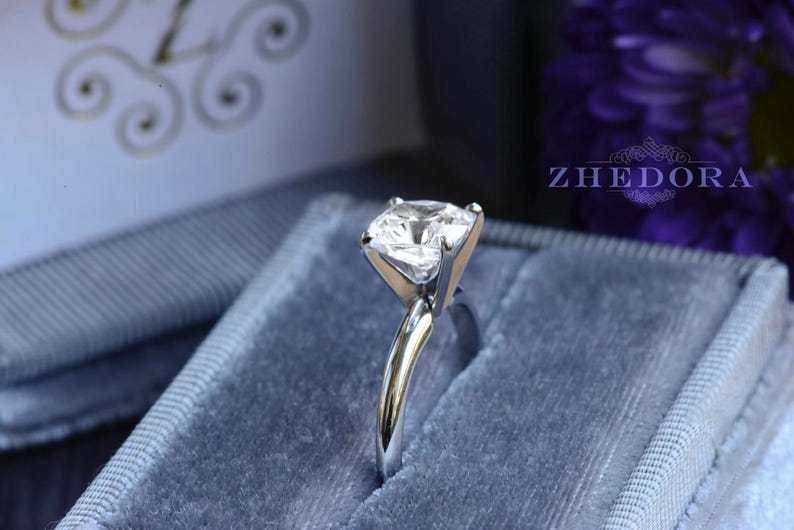May include: A silver ring with a clear, square-shaped gemstone in a blue velvet ring box. The box has the word "ZHEDORA" printed on it.