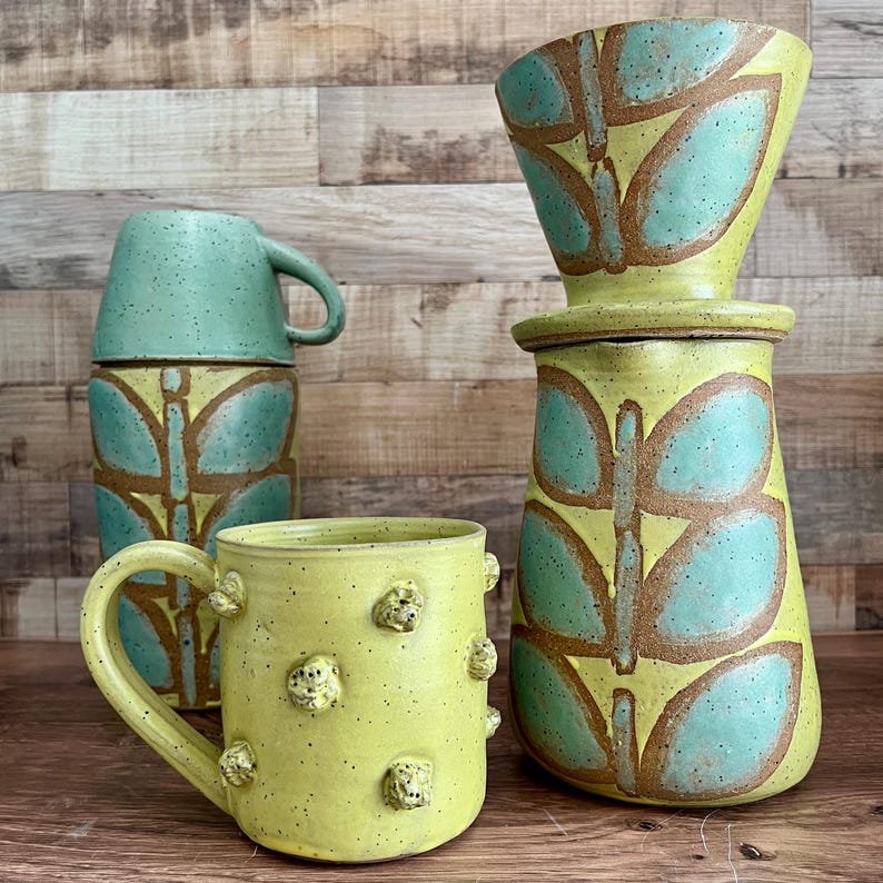 May include: A collection of handmade ceramic kitchenware. The set includes a yellow mug with textured details, a turquoise mug, and a yellow coffee maker with a lid. All pieces feature a butterfly design in turquoise and brown.