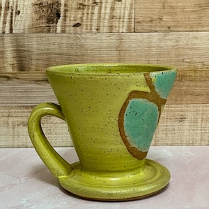May include: A handmade ceramic pour-over coffee maker in a pale yellow color with a matching handle and base. The coffee maker has a unique design with brown and teal accents. The speckled surface adds to its rustic charm.