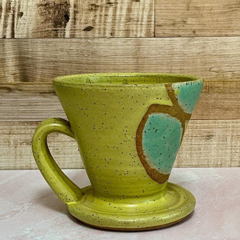 May include: A handmade ceramic pour-over coffee maker in a pale yellow color with a matching handle and base. The coffee maker has a unique design with brown and teal accents. The speckled surface adds to its rustic charm.