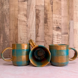 May include: A set of three ceramic mugs with a speckled, matte finish. The mugs are in shades of cream, light gray, and dark gray. Each mug has a simple, minimalist design, perfect for coffee or tea.