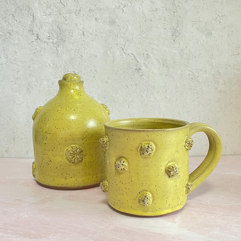 May include: A yellow ceramic mug and a matching bottle with a lid. Both pieces have a speckled finish and are adorned with raised, textured floral designs. The mug has a handle, and the bottle has a rounded shape.