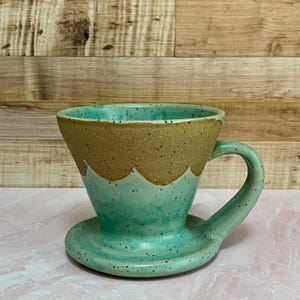 May include: A handmade ceramic pour-over coffee maker in shades of turquoise and tan. The coffee maker has a handle and a matching saucer. The top rim is tan with a speckled texture, and the bottom is turquoise with a speckled finish.