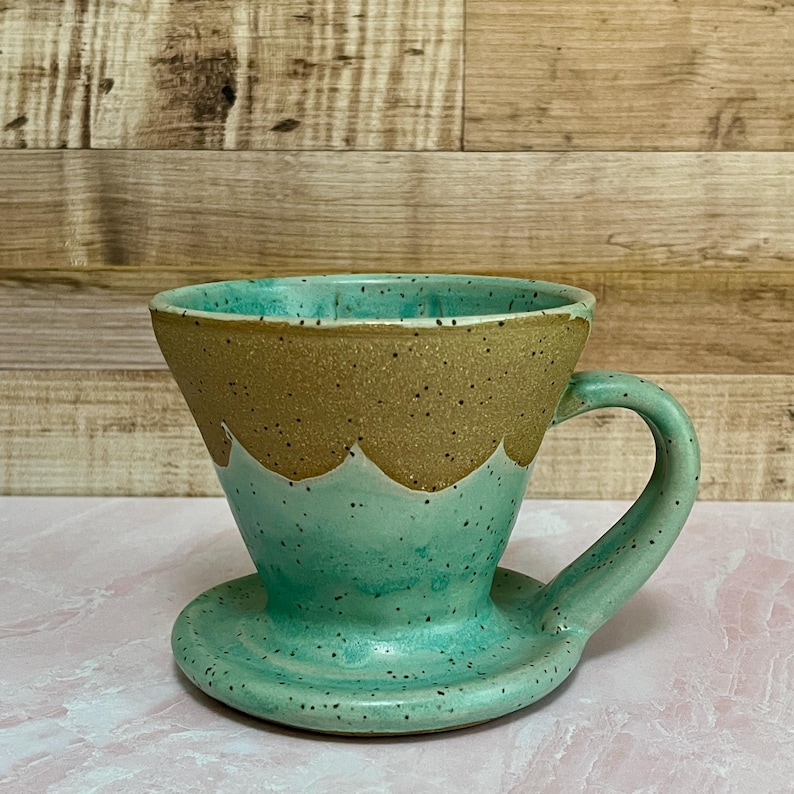 May include: A handmade ceramic pour-over coffee maker in shades of turquoise and tan. The coffee maker has a handle and a matching saucer. The top rim is tan with a speckled texture, and the bottom is turquoise with a speckled finish.