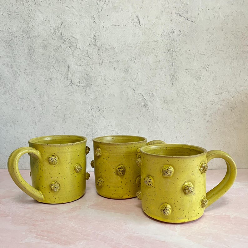 May include: Three handmade ceramic mugs in a vibrant yellow hue. Each mug features a speckled texture and is adorned with small, raised, textured embellishments. The mugs have curved handles and are displayed on a pink surface against a neutral background.