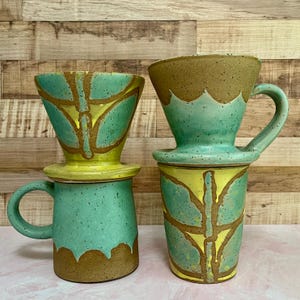 May include: Two ceramic pour-over coffee makers. One is a mug with a handle, the other is a tall cup. Both are turquoise with yellow and brown leaf-like designs. The coffee makers have a speckled texture and a rustic aesthetic.