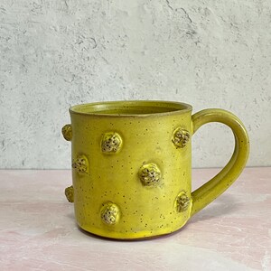 May include: A handmade yellow ceramic mug with a large handle and textured, raised, brown, and yellow bumps. The mug has a speckled finish and is set against a pink and gray background. This unique mug is perfect for coffee or tea.