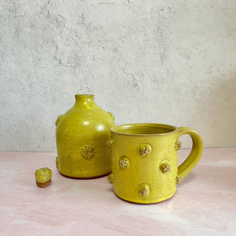 May include: A yellow ceramic mug and vase set with textured, raised details. The mug has a handle and the vase is a rounded shape. The set is speckled with dark spots and has a matching stopper.