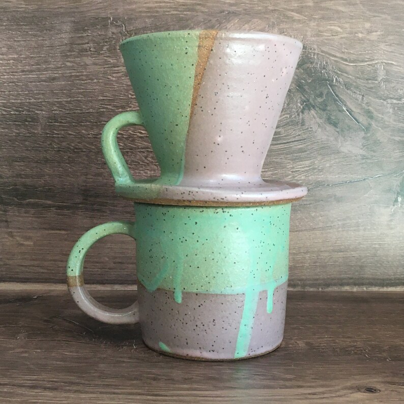 May include: A ceramic coffee pour-over set with a mug and a dripper. The mug and dripper are divided into two color sections: mint green and lavender. The mug has a handle with a gold accent. The dripper sits atop the mug.