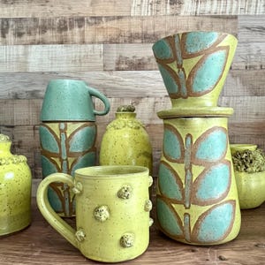 May include: A collection of handmade ceramic pottery in shades of yellow and teal. The pieces include mugs, a coffee dripper, and decorative jars, all featuring a butterfly design. The pottery has a speckled texture and a rustic aesthetic.