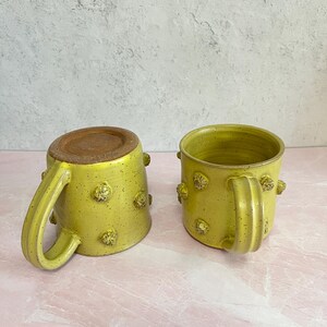 May include: Two handmade ceramic mugs. The mugs are a speckled yellow-green color with textured, raised, decorative elements. Each mug has a handle and a brown interior base. The mugs are on a pink surface.