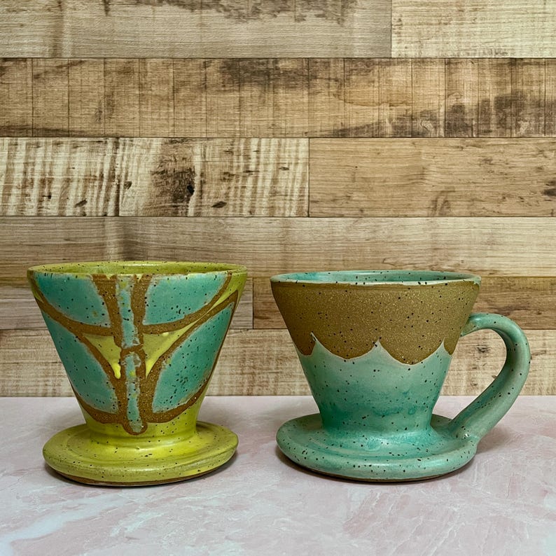 May include: Two ceramic pour-over coffee makers. One is yellow and teal with a brown design. The other is teal and brown with a handle. Both have a circular base and a cone shape.