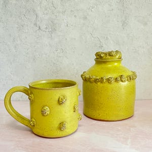May include: A yellow ceramic mug and a jar with a lid. Both pieces have a speckled finish and are adorned with small, textured, raised details. The mug has a handle, and the jar has a decorative rim around the top.