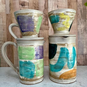 May include: Two ceramic coffee pour-over sets. Each set includes a pour-over cone and a carafe. The sets are painted with colorful abstract designs in shades of green, purple, blue, and yellow. The sets are handmade.