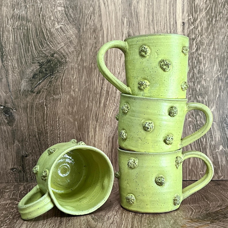 May include: Four stacked, handmade ceramic mugs in a vibrant lime green. Each mug features a large, curved handle and is adorned with textured, raised, circular embellishments. The mugs are displayed against a wooden background.