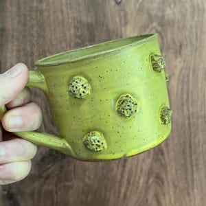 May include: A handmade ceramic mug in a vibrant chartreuse green. The mug features textured, raised, organic-shaped embellishments and a speckled finish. The handle is curved, and the mug is set against a wooden background.