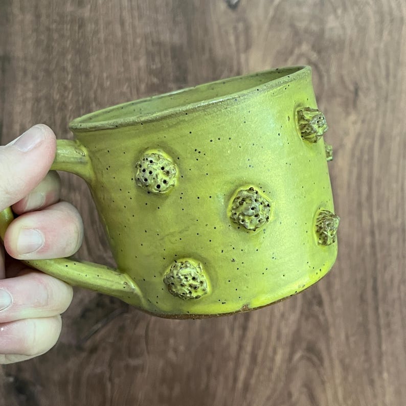 May include: A handmade ceramic mug in a vibrant chartreuse green. The mug features textured, raised, organic-shaped embellishments and a speckled finish. The handle is curved, and the mug is set against a wooden background.