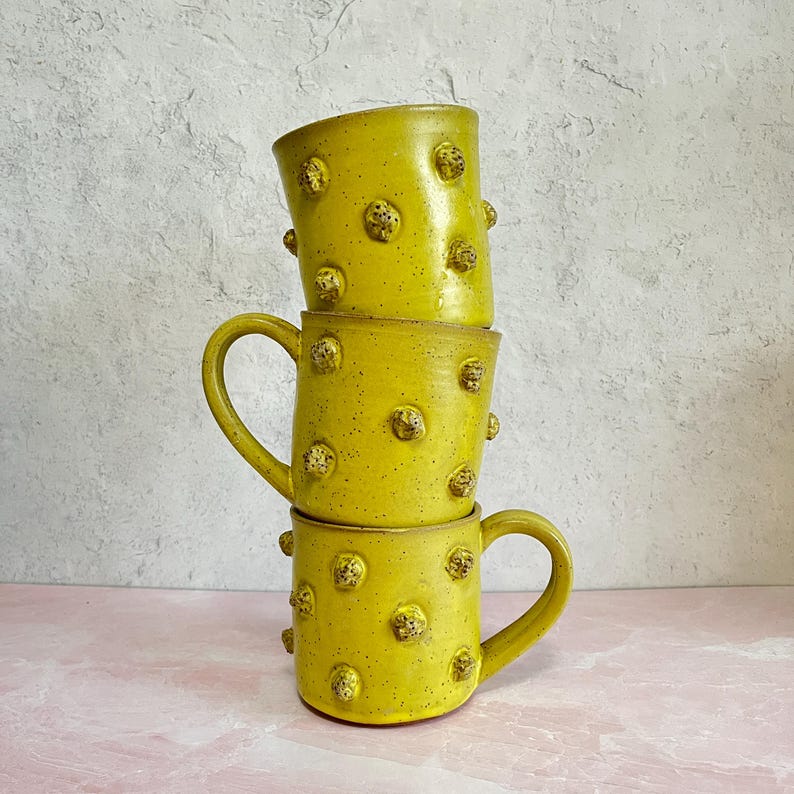May include: Three stacked, handmade ceramic mugs. The mugs are a vibrant yellow with a textured surface and small, raised, rounded embellishments. Each mug has a curved handle. The mugs are stacked on top of each other.