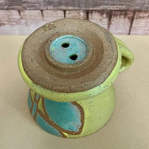 May include: A handmade ceramic mug with a unique design. The mug features a light green base with turquoise and brown accents. The top has a brown rim and a turquoise interior with two holes. The handle is a matching light green color.