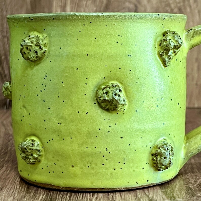 May include: A handmade ceramic mug in a vibrant lime green color, speckled with black dots. The mug features six textured, raised, organic-shaped embellishments. It has a curved handle and a slightly uneven rim, showcasing its handcrafted nature.