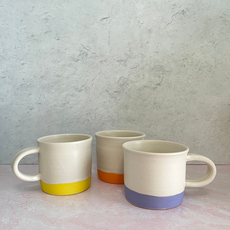 A cheery white ceramic coffee mug with an orange, yellow, or lilac bottom stripe. This was handmade by me. I threw it on a pottery wheel, and painted an underglaze stripe on the bottom, then fired it with a white glaze.